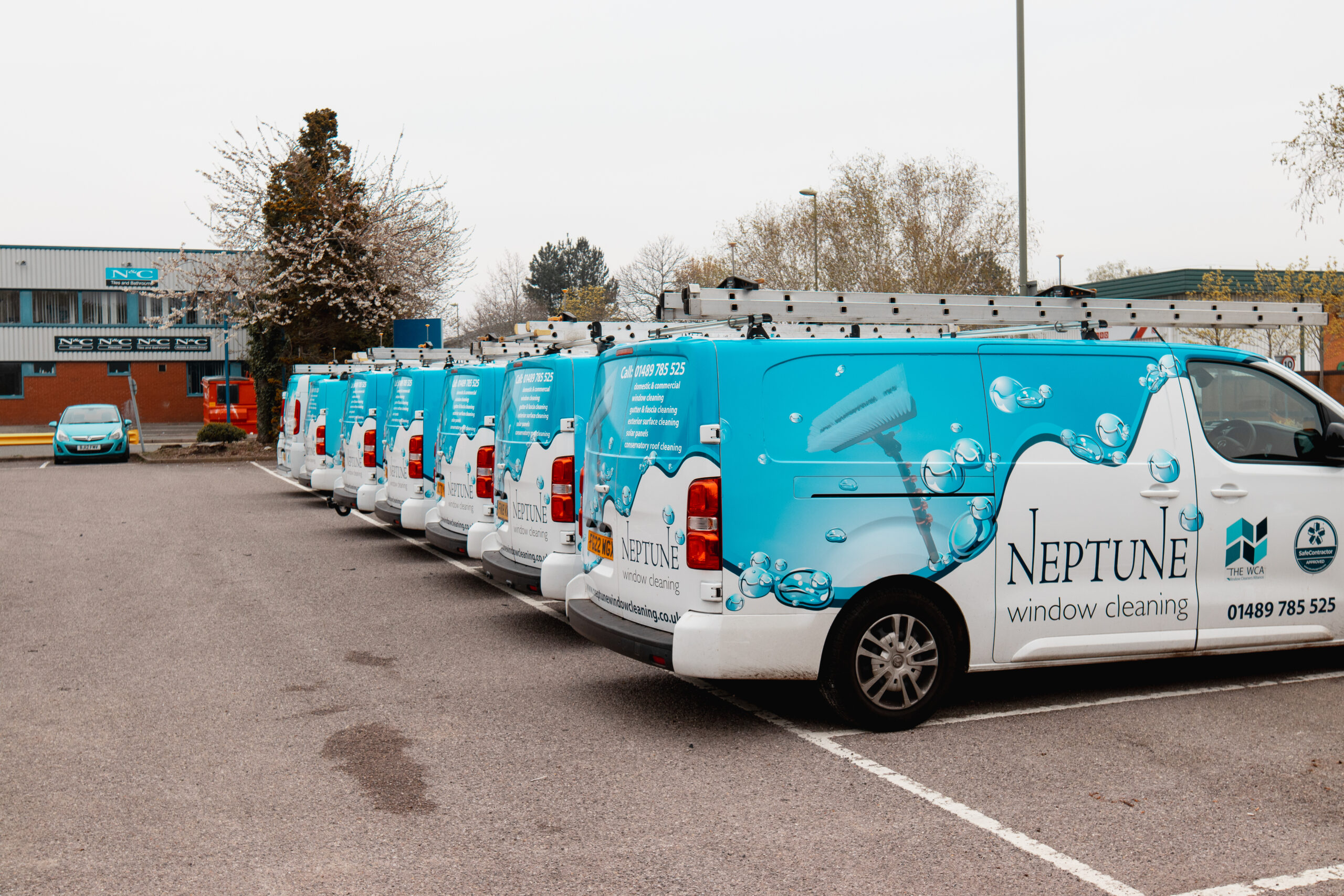 A row of Neptune Window Cleaning branded vans parked in a car park, each fitted with roof ladders and displaying the company’s blue and white livery, representing their professional window cleaning fleet.