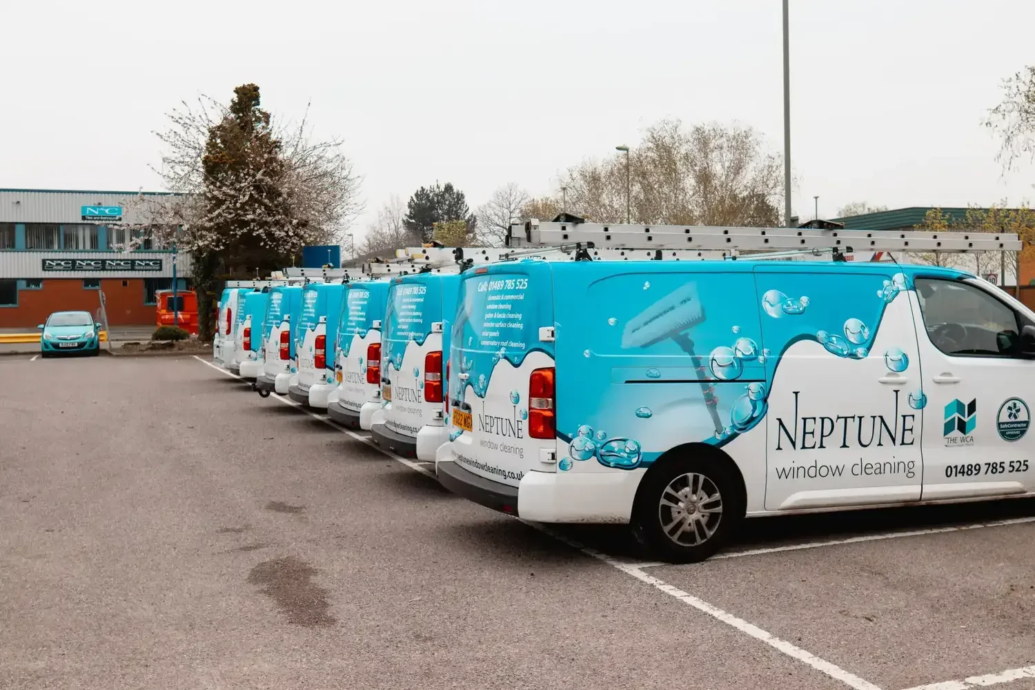 A row of Neptune Window Cleaning branded vans parked in a car park, each fitted with roof ladders and displaying the company’s blue and white livery, representing their professional window cleaning fleet.