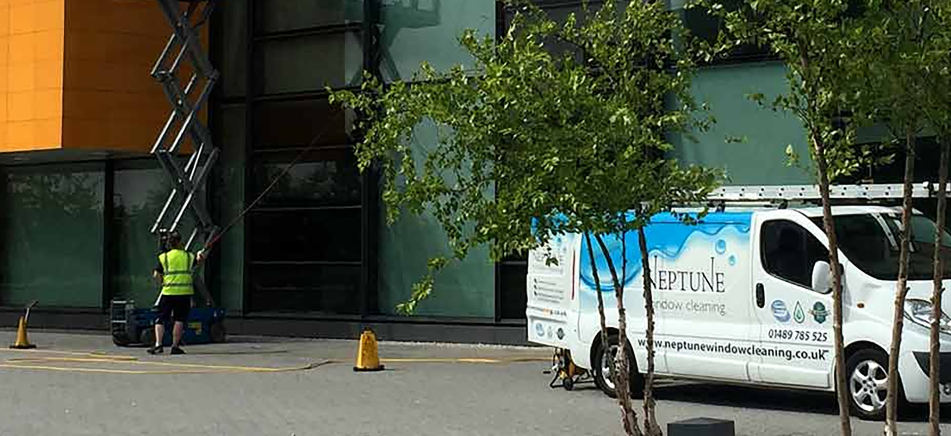 A Neptune Window Cleaning van parked outside a modern commercial building while a technician in high-visibility clothing operates a lift to clean upper-floor windows, demonstrating professional commercial window cleaning services.